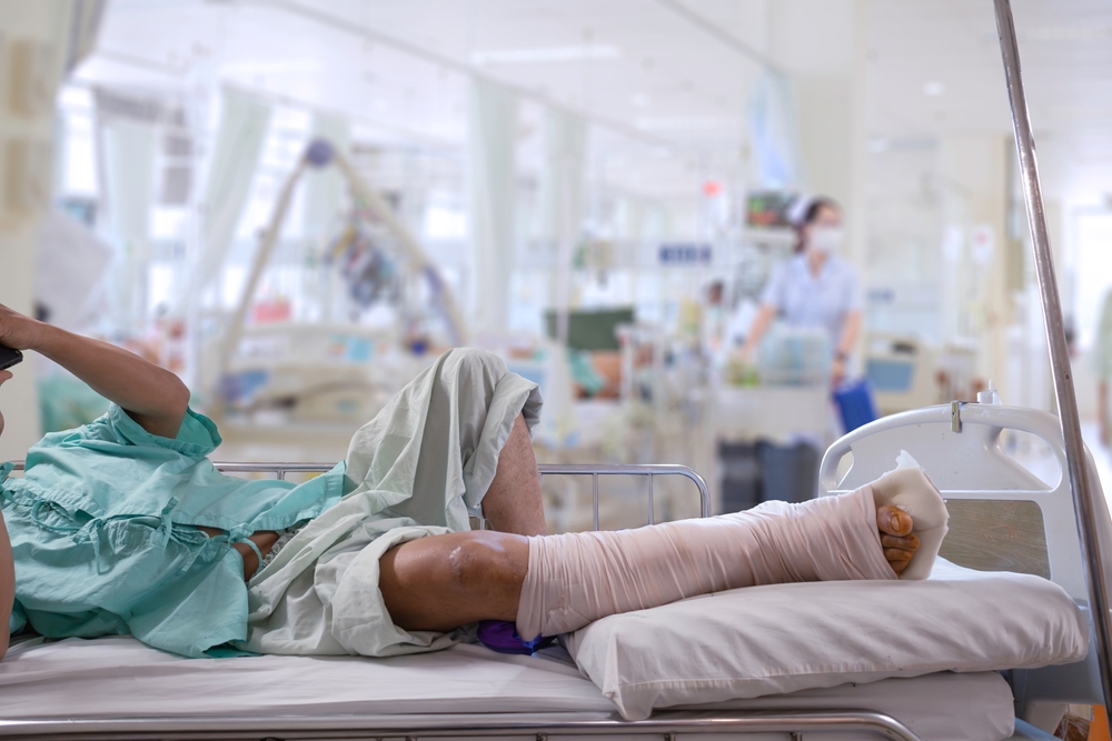 A patient with a broken leg in a cast lies in a hospital bed with a nurse taking care of him.