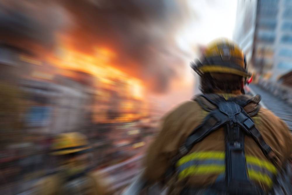 A firefighter in full gear, seen from behind, moves towards a raging inferno engulfing a building.